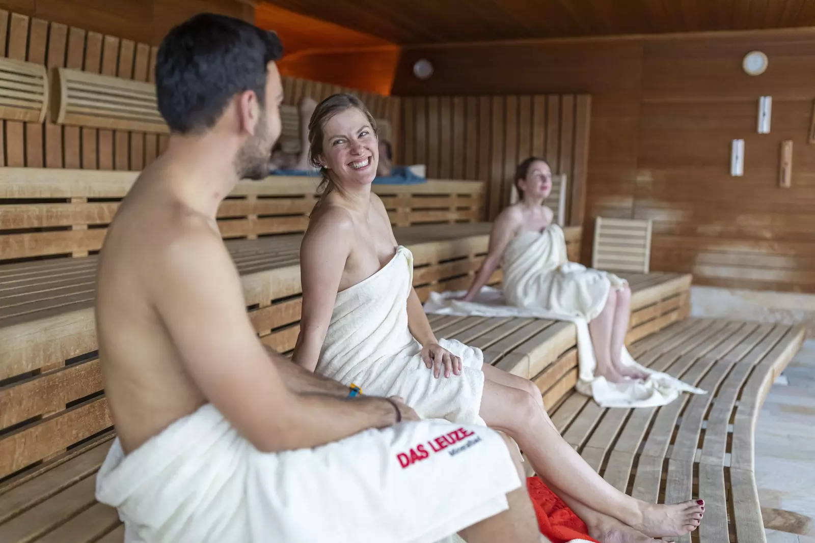 Ein Paar, in Badetücher gehüllt sitzt auf einer Holzbank in der Panorama-Sauna und unterhält sich entspannt und fröhlich.