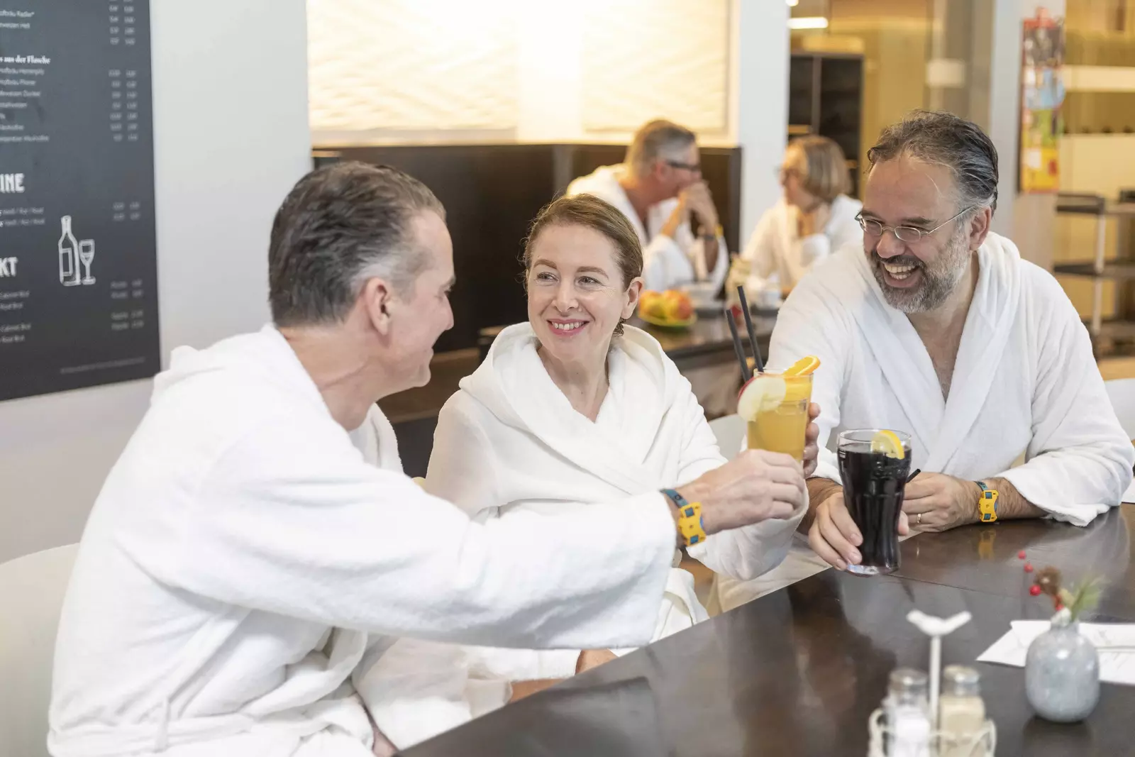 Zwei Männer und eine Frau in weißen Bademänteln genießen kalte Getränke an der Theke im Restaurant im LEUZE.