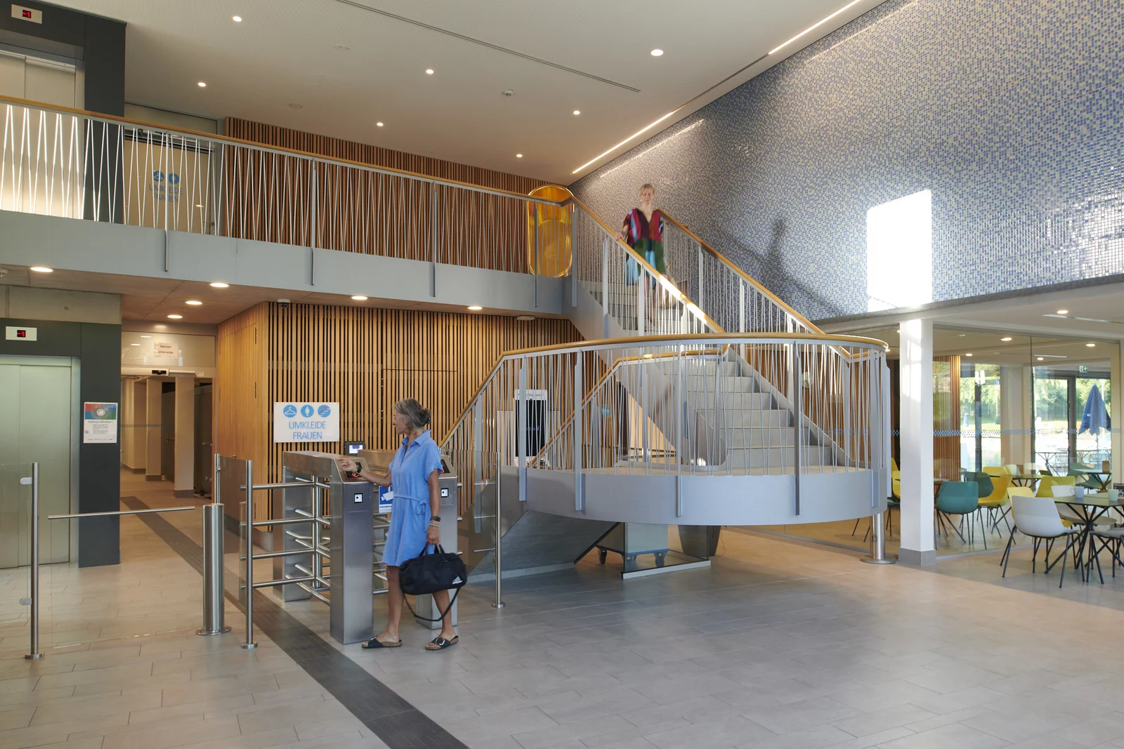 Eingangsbereich ins Bad im Foyer Mineralbad Berg Eine Frau geht durch die Schranke im Foyer ins Bad. Eine weitere Frau schreitet die Treppe, vorbei an der goldenen Badewanne hinunter.