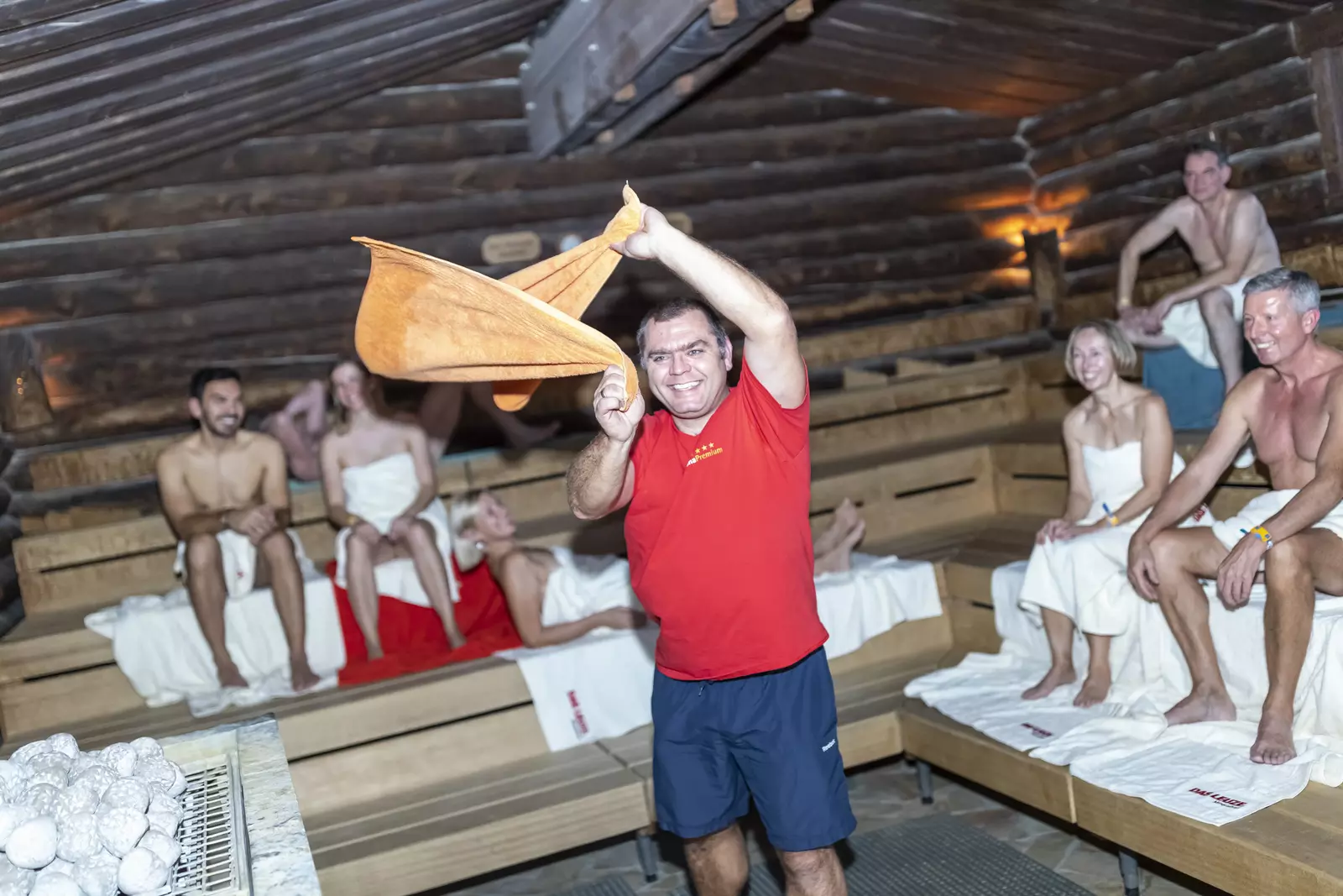 Saunabesucher genießen fröhlich die Aufguss-Zeremonie in der Kelo-Sauna im LEUZE. Der Aufgussleiter wirbelt mit einem roten Handtuch die heiße Saunaluft auf.