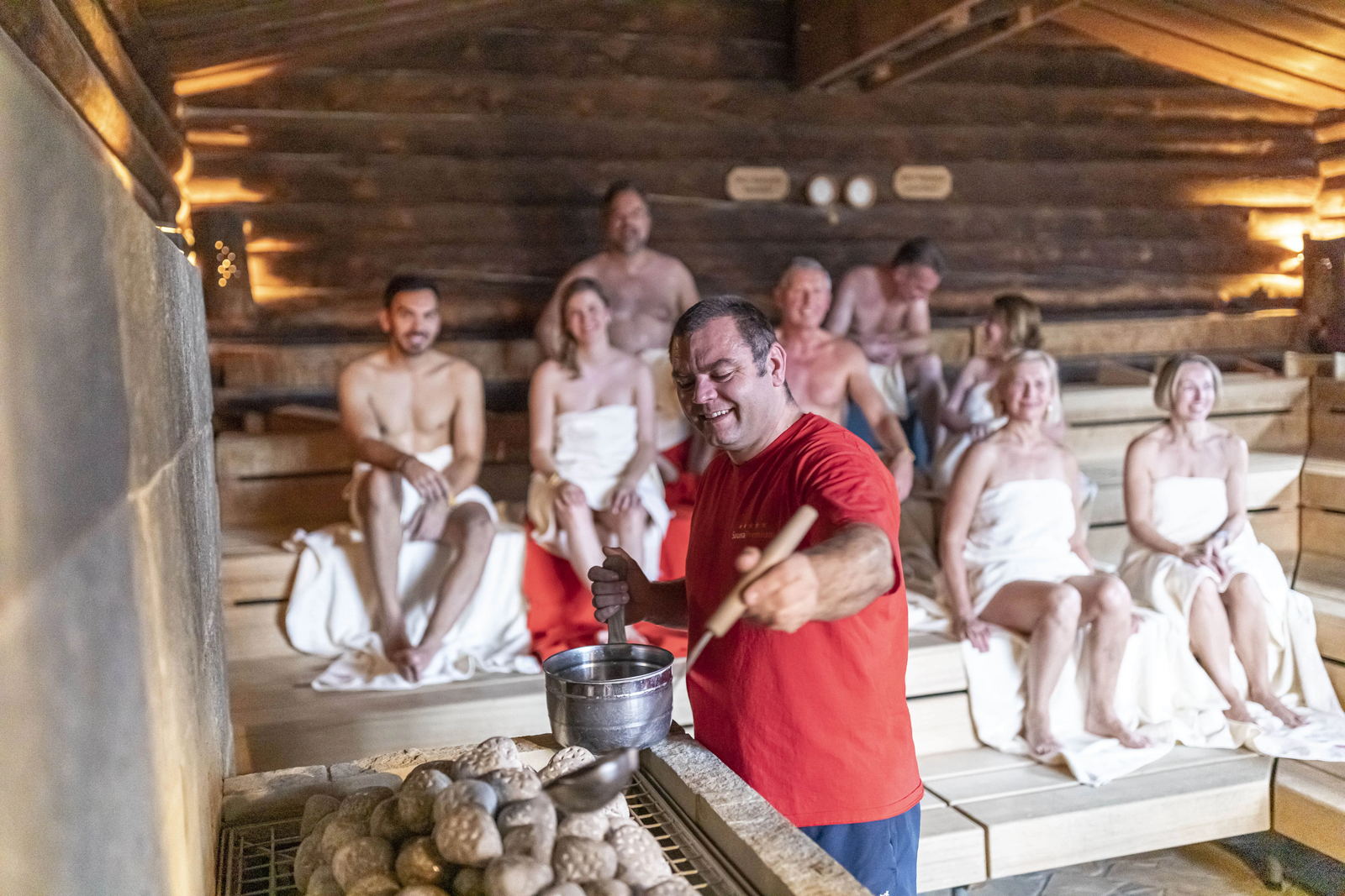 Fröhliche Saunagäste sitzen auf den Holzbänken in der Sauna im LEUZE. Ein gutgelaunter Saunameister bereitet einen Aufguss vor.