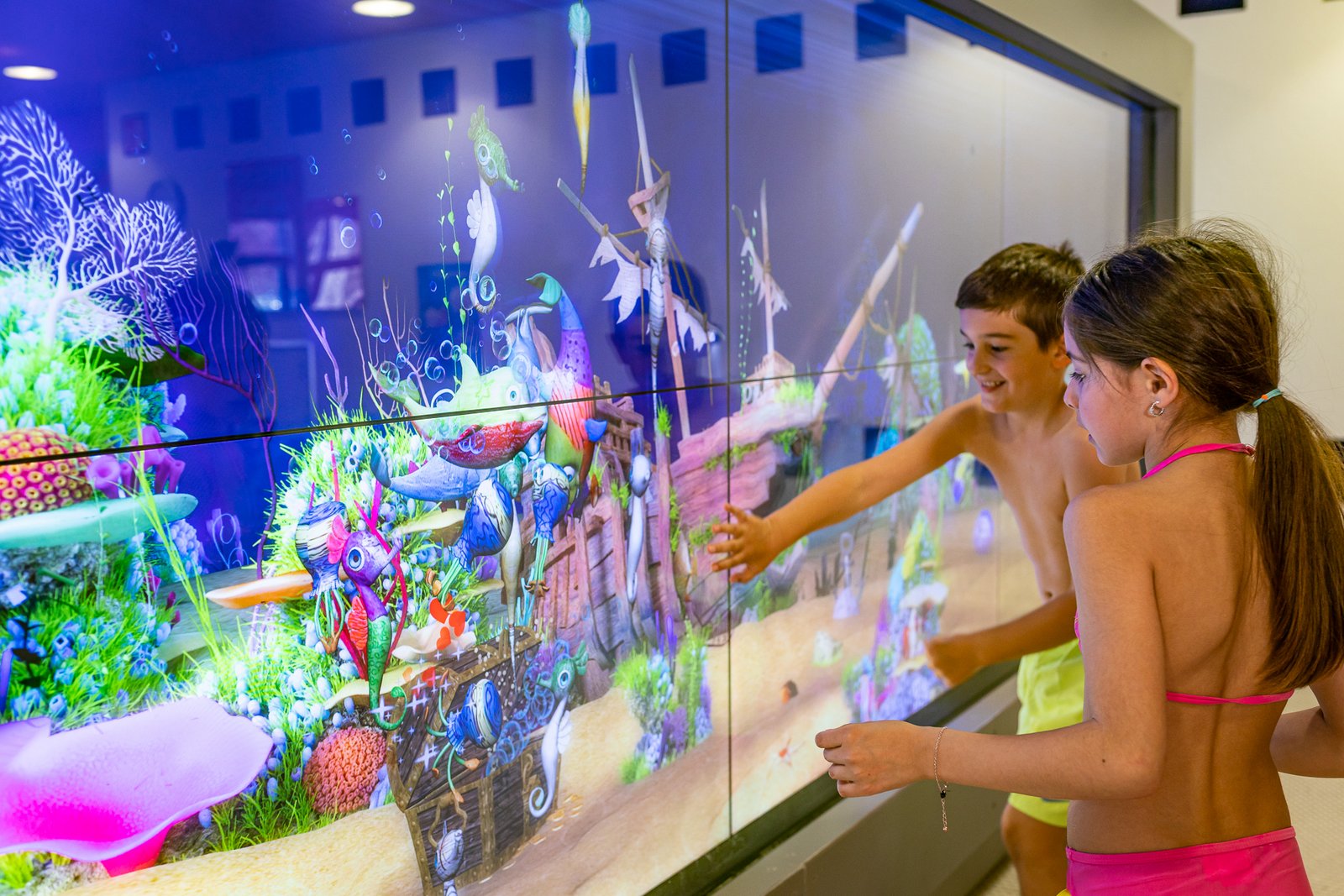 An der Wand bunte Unterwasserwelt des interaktiven Aquariums.. Ein Junge und ein Mädchen davor und spielen über Touch-Funktion mit den Wassertieren.