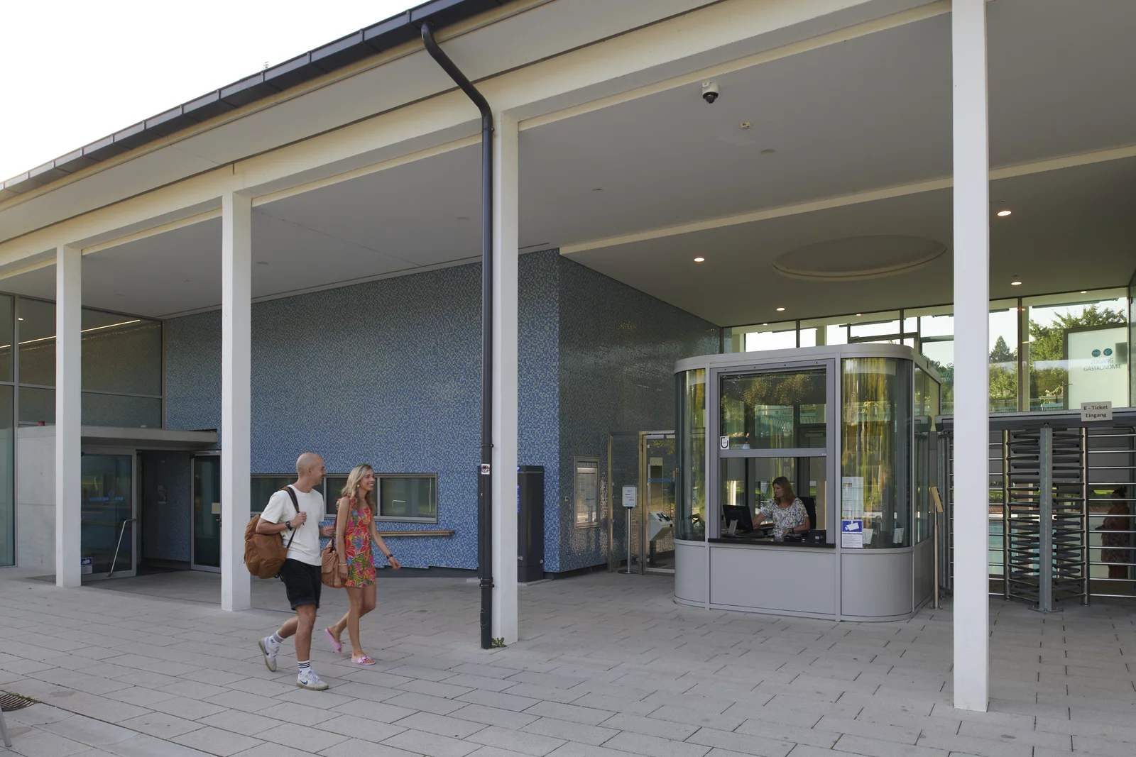 Eingang außen ins Mineralbad Berg Ein jünges Paar läuft Richtung Kasse zum Eingang mit dem Drehkreuz an der Badfassade entlang.