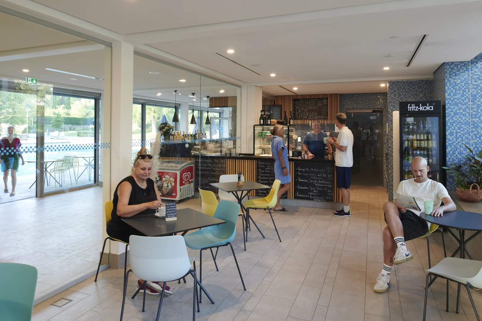 Café im Mineralbad Berg Gemütliches Café im Mineralbad Berg. Eine Frau sitzt am Tisch links im Bild. Rechts am Tisch liest ein jünger Mann eine Zeitschrift. Im Hintergrund ist eine Verkaufstheke zu sehen.