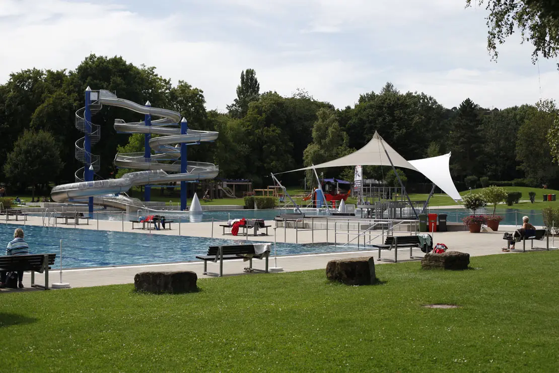 Außenansicht Beckenlandschaft mit Liegewiese, großer Wasserrutsche und Sitzbänken im Freibad Rosental.
