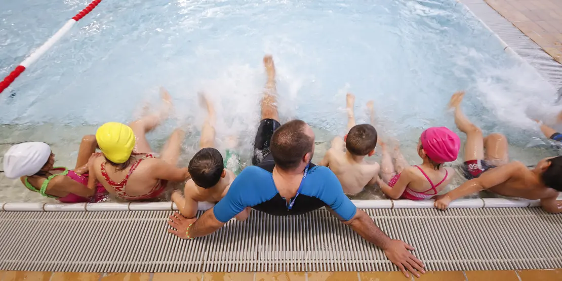 Rückenansicht Kinder in Begleitung vom Schwimmkursleiter am Beckenrand im Wasser während des Kinderschwimmkurs im Hallenbad Sonnenberg.